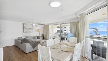 A modern living room with a sofa, dining table, and chairs, a balcony with a view of the sea, and a ceiling light.