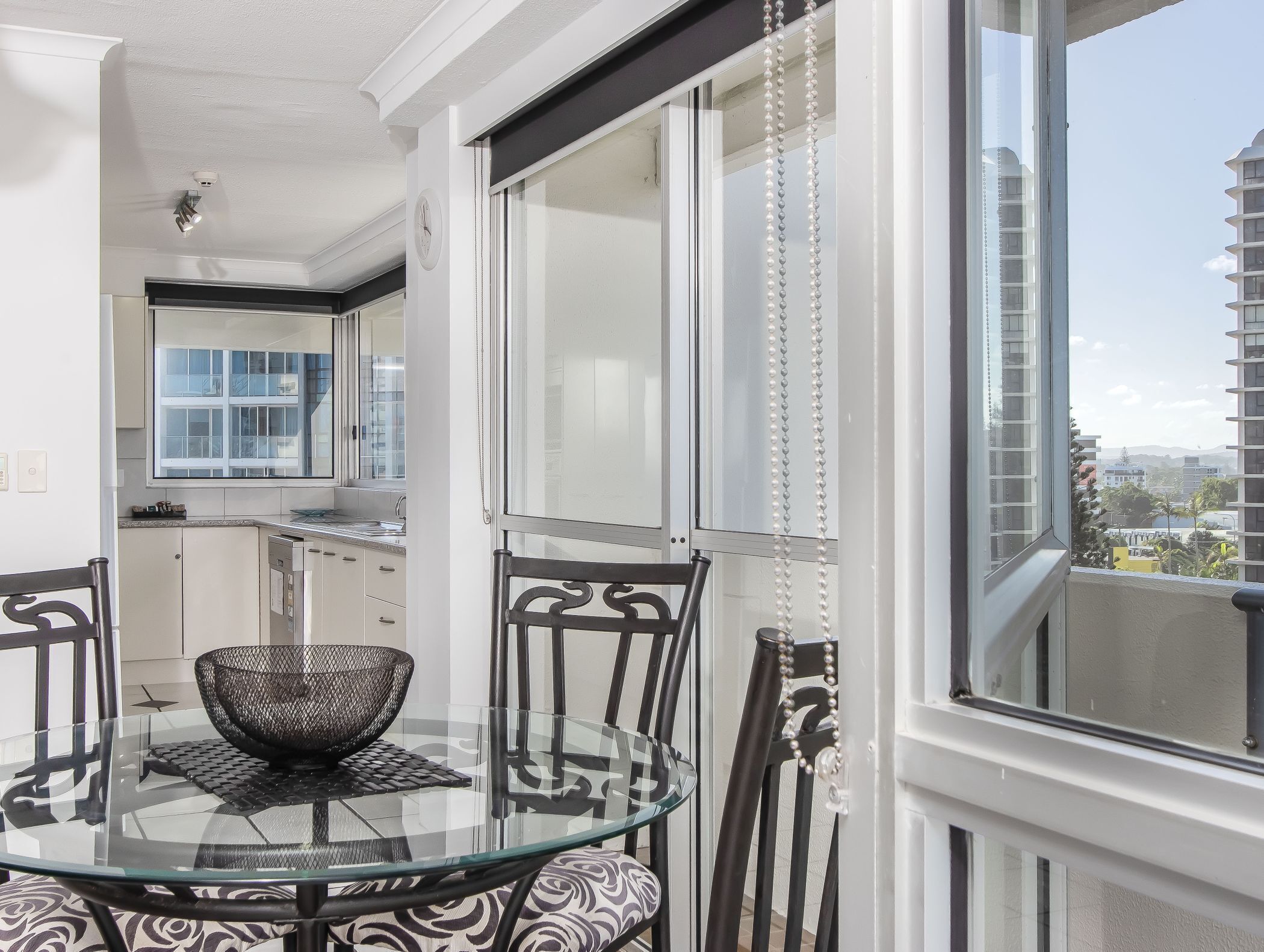 A modern kitchen with a window, a dining area with a glass table and chairs, and a view of the cityscape.
