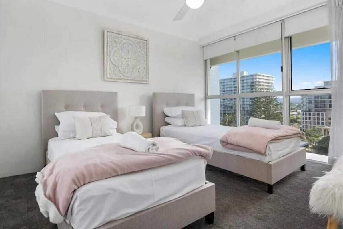Una habitación de hotel moderna con dos camas, un ventilador de techo, un ventanal con vistas a la ciudad y un elemento decorativo en la pared.