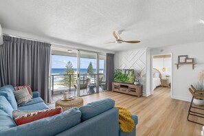 A modern living room with a blue sofa, a ceiling fan, a large window with a view of the ocean, and a wooden floor. - Baronnet Apartments (Surfers Paradise)