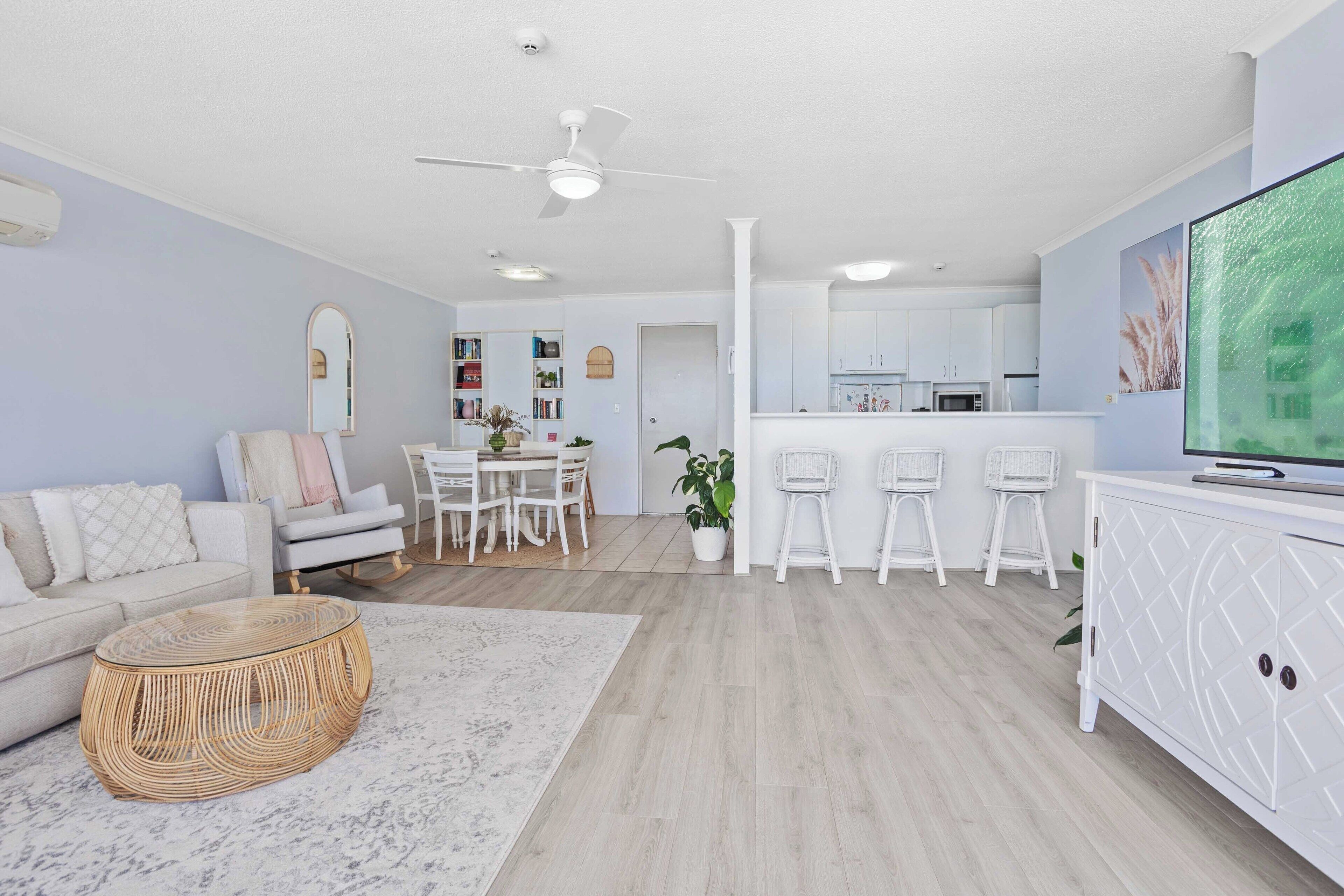 Una sala moderna con sofá, mesa de centro y televisor. Se observa un comedor con sillas y una mesa, y una cocina al fondo.