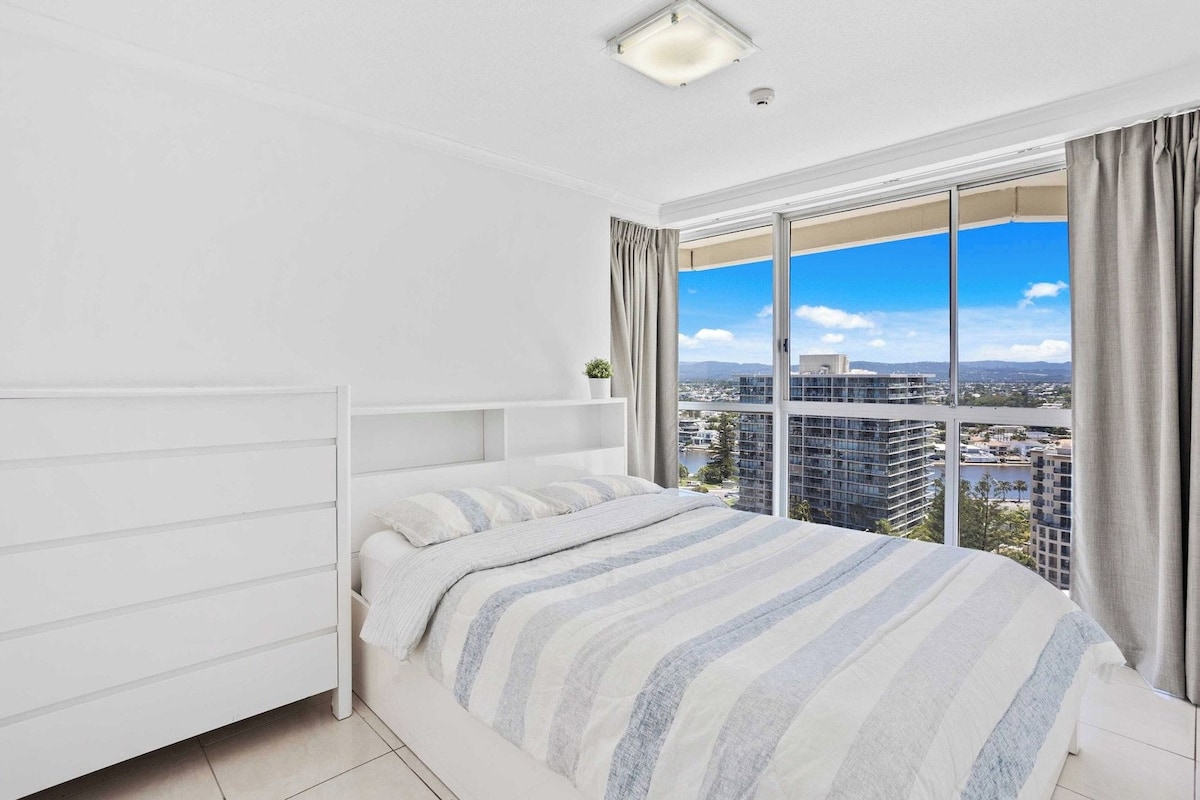 Un dormitorio moderno con una cama grande, un cabecero blanco y vistas a un paisaje urbano a través de amplias ventanas.