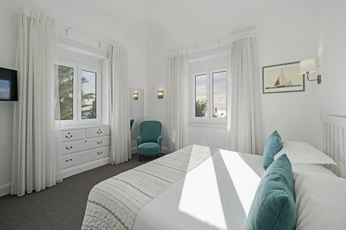 Une chambre d’hôtel avec un grand lit, une chaise, une commode et deux fenêtres avec des rideaux.