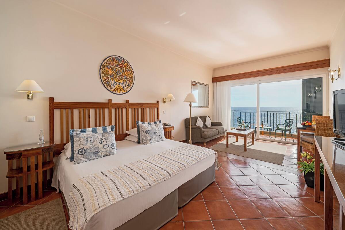 Une chambre spacieuse avec un grand lit, une tête de lit en bois, un coin repas avec une table et des chaises, et un balcon avec vue.