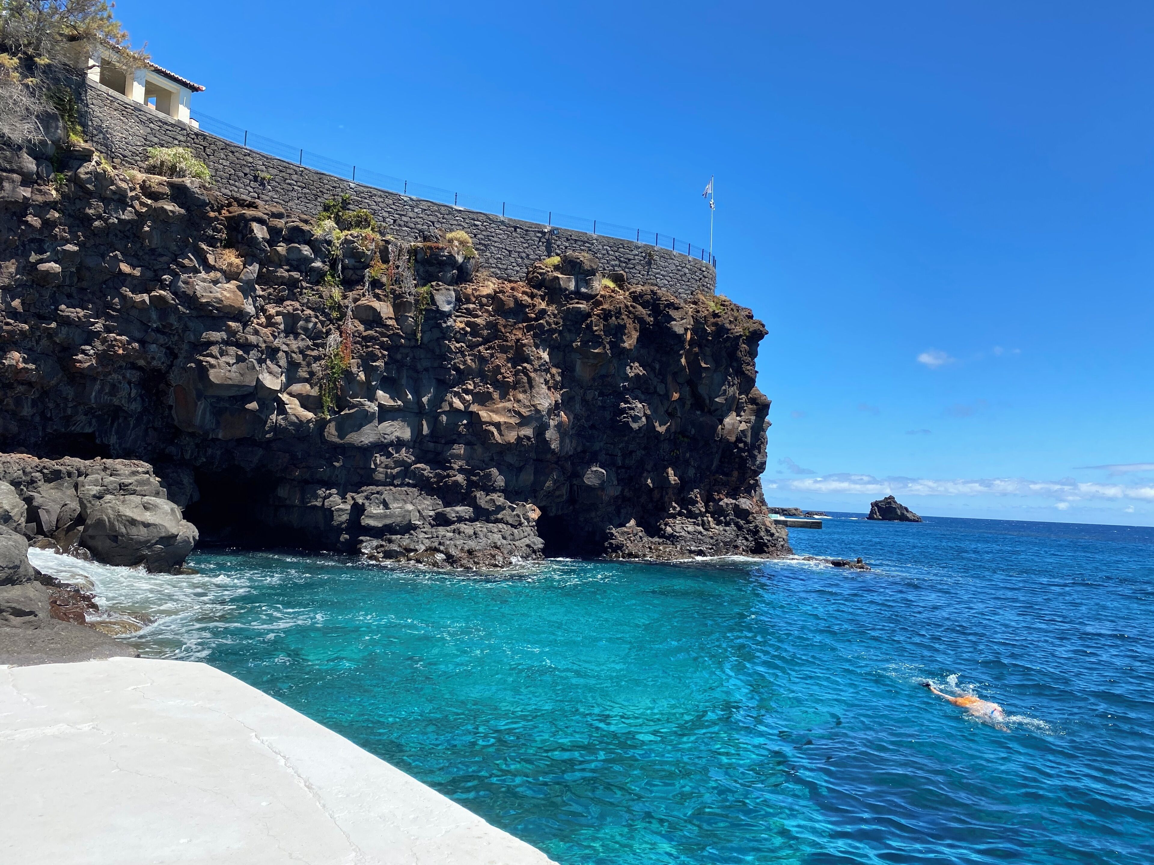 A coastal scene with a rocky cliff, clear blue water, and a swimmer.