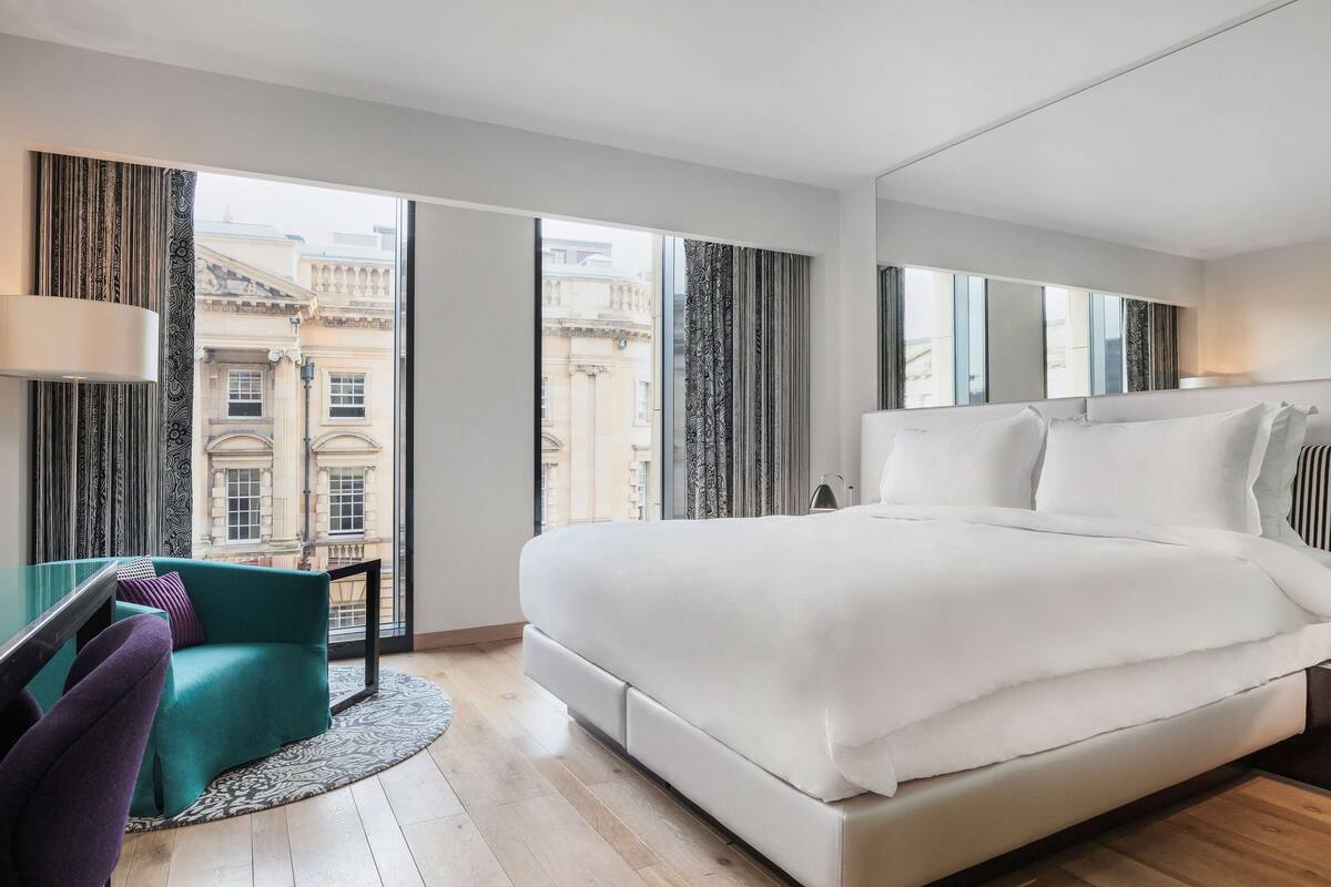 Une chambre d’hôtel moderne dotée d’un grand lit, d’un coin salon avec un fauteuil de couleur turquoise et offrant une vue sur des bâtiments historiques à travers de grandes fenêtres.