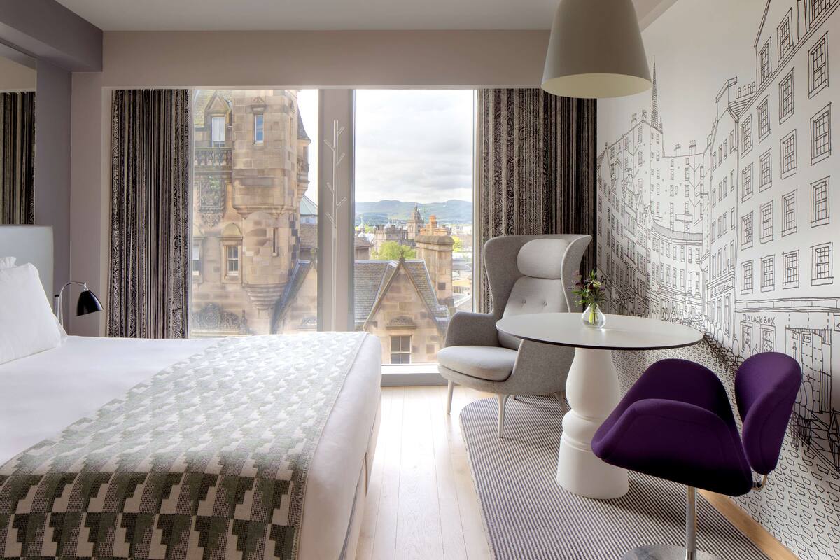 Une chambre d’hôtel avec un grand lit, une table à manger, deux chaises et une vue sur le paysage urbain, visible à travers de grandes fenêtres.