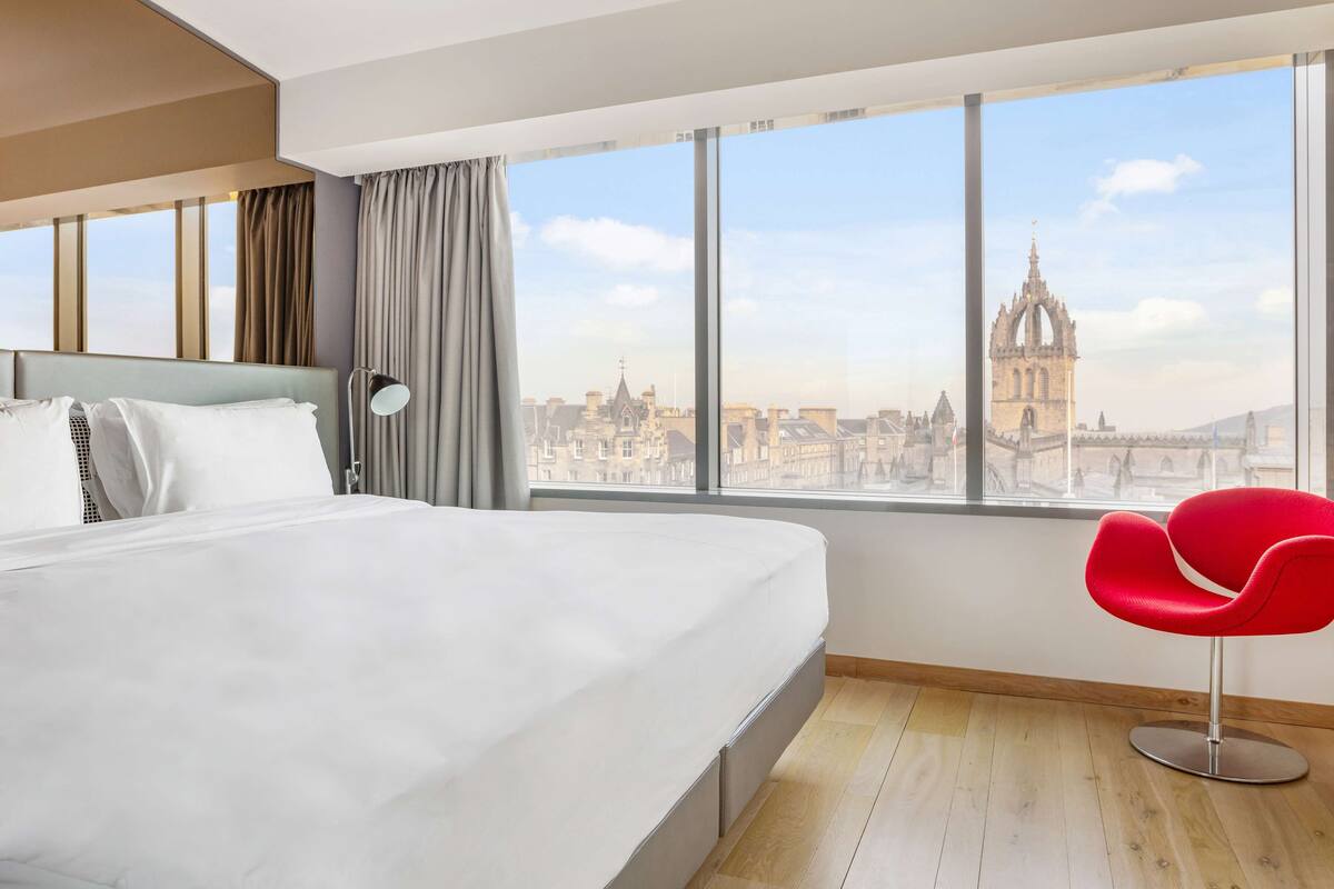 Une chambre d’hôtel moderne avec un grand lit, un fauteuil rouge et une vue sur un bâtiment historique.