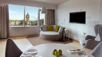A modern living room with a round table, grey armchairs, a sofa with cushions, a TV mounted on the wall, and a view of a cityscape through large windows.