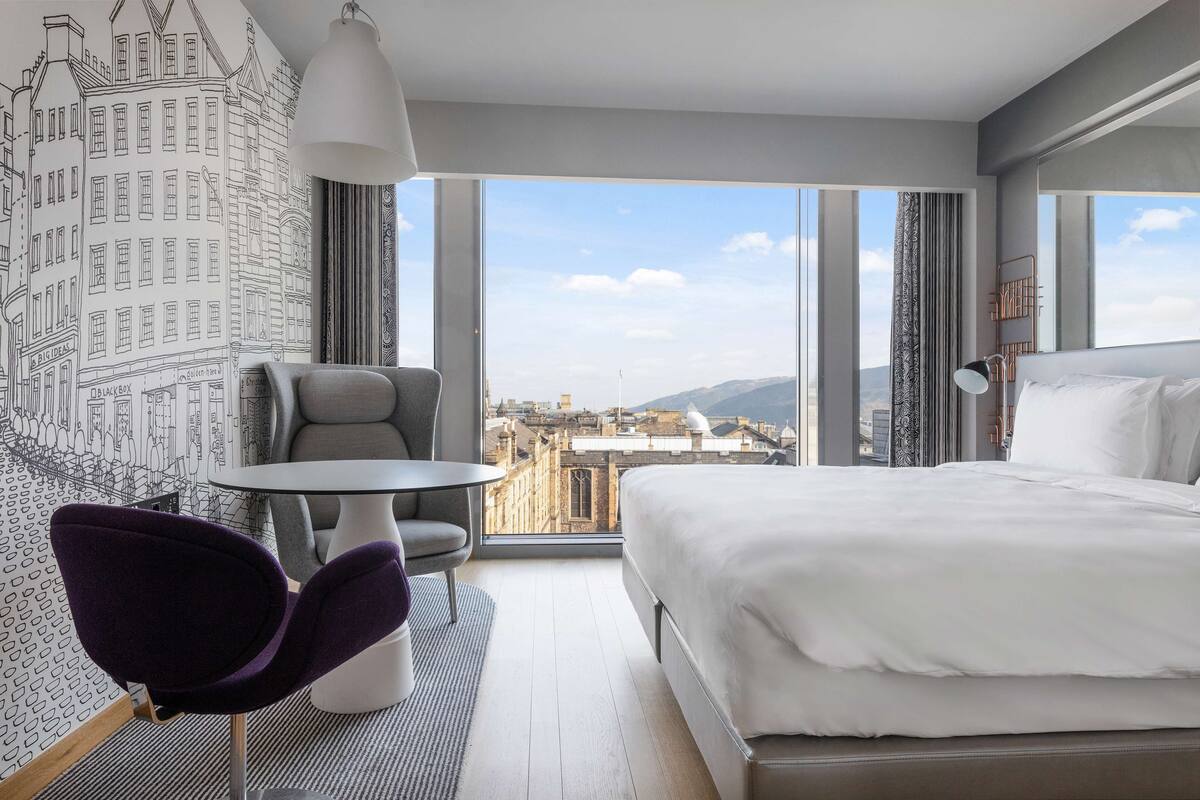 Une chambre d’hôtel avec un grand lit, une chaise, une petite table et une vue sur le paysage urbain grâce à de grandes fenêtres.