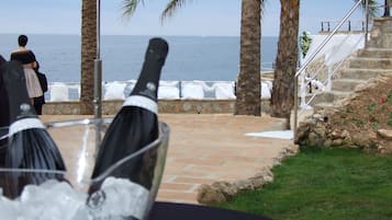 Una mesa con una botella de champán, una copa y una botella de agua. Se ven palmeras y una escalera de piedra que conduce a un balcón con vistas al mar.