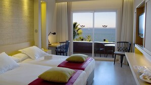 A hotel room with two beds, a balcony with a view of the sea, a desk, and a TV.