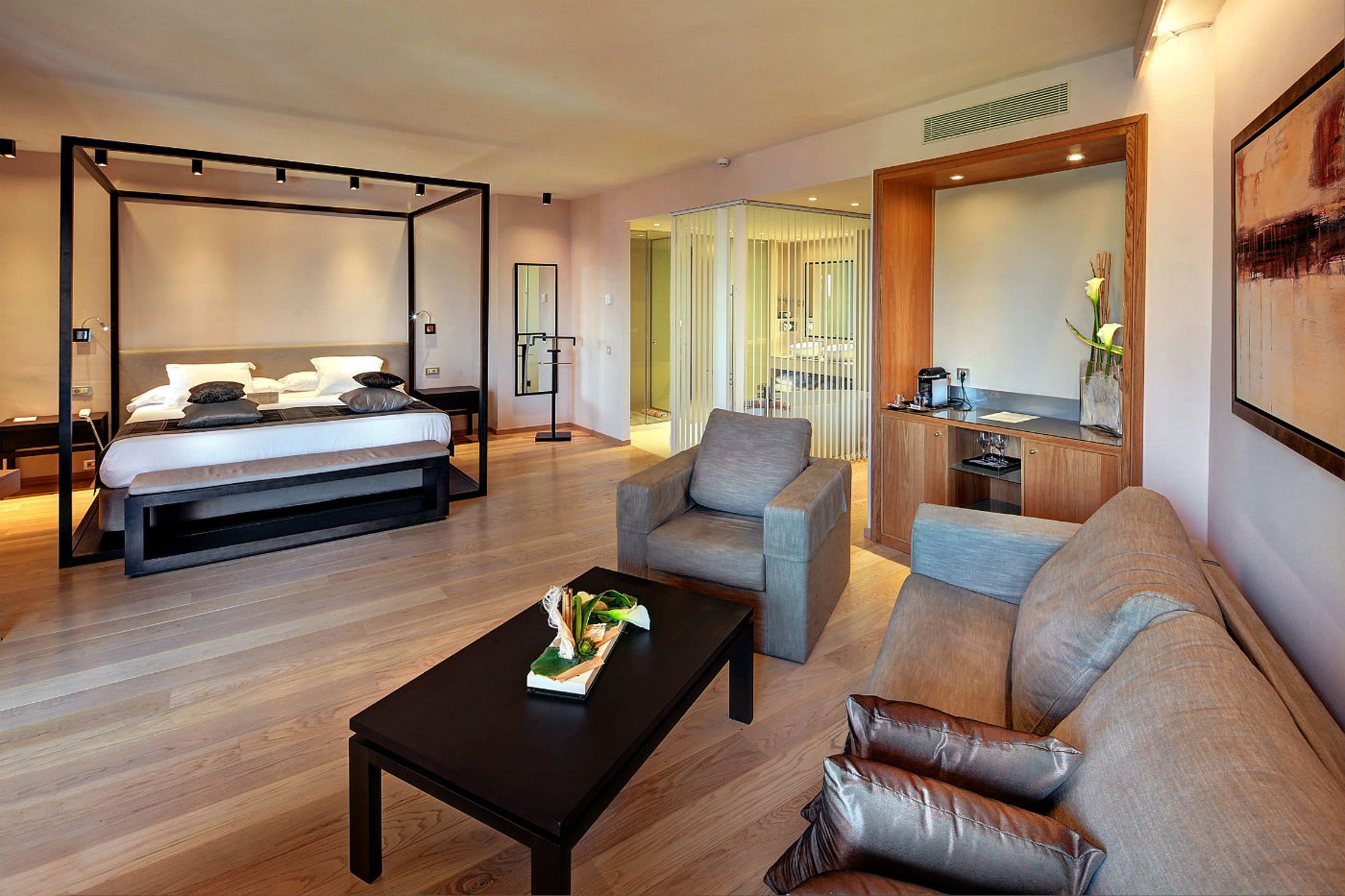 A modern hotel room with a four-poster bed, a gray armchair, a brown leather sofa, and a coffee table.