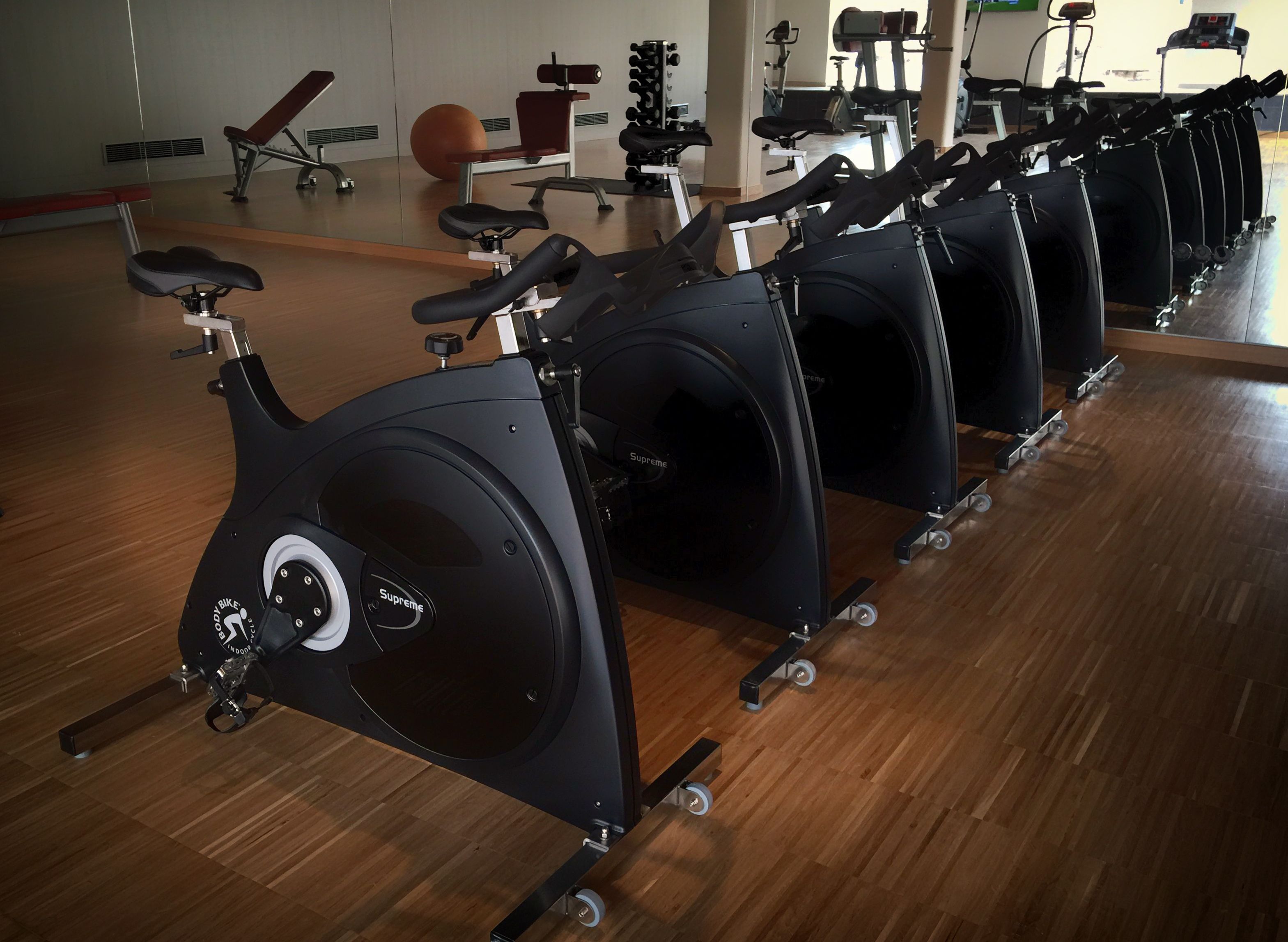 Varias bicicletas estáticas en un gimnasio con suelo de madera y espejos en las paredes.