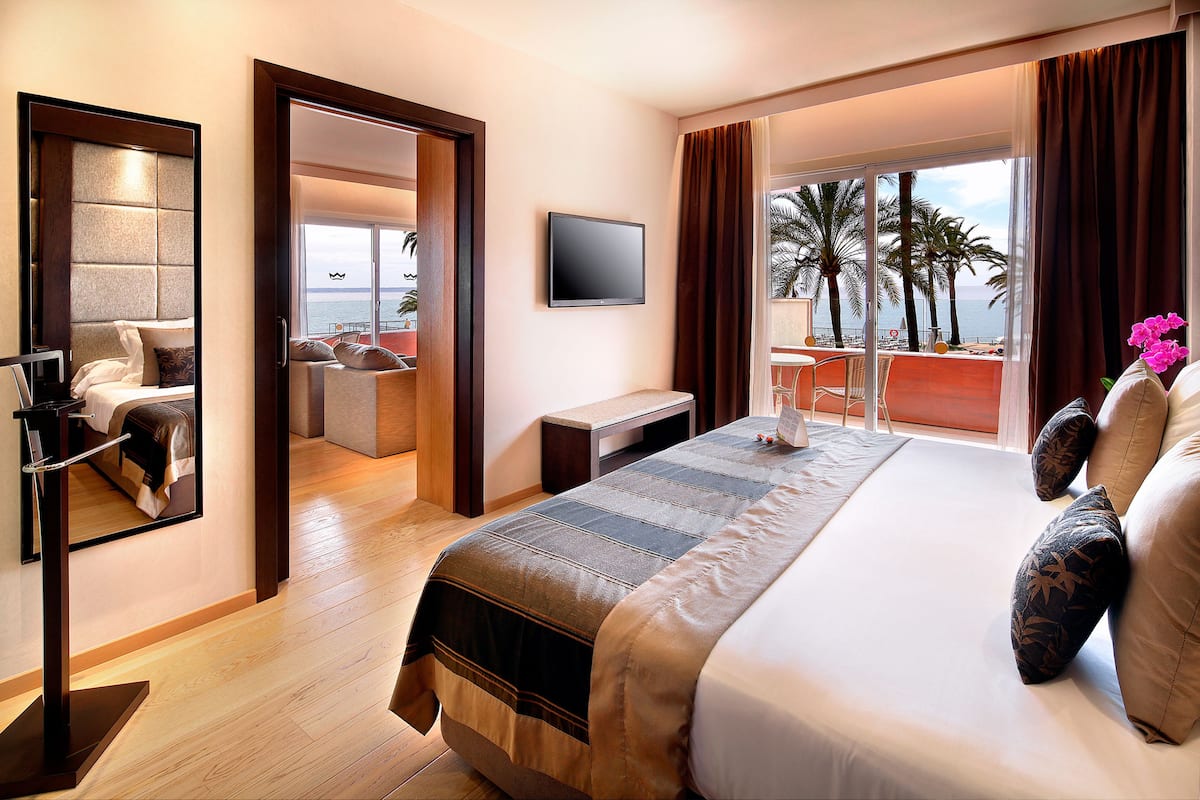 Une chambre d’hôtel dotée d’un grand lit, d’une télévision, d’un bureau et d’un balcon offrant une vue sur les palmiers et la mer.