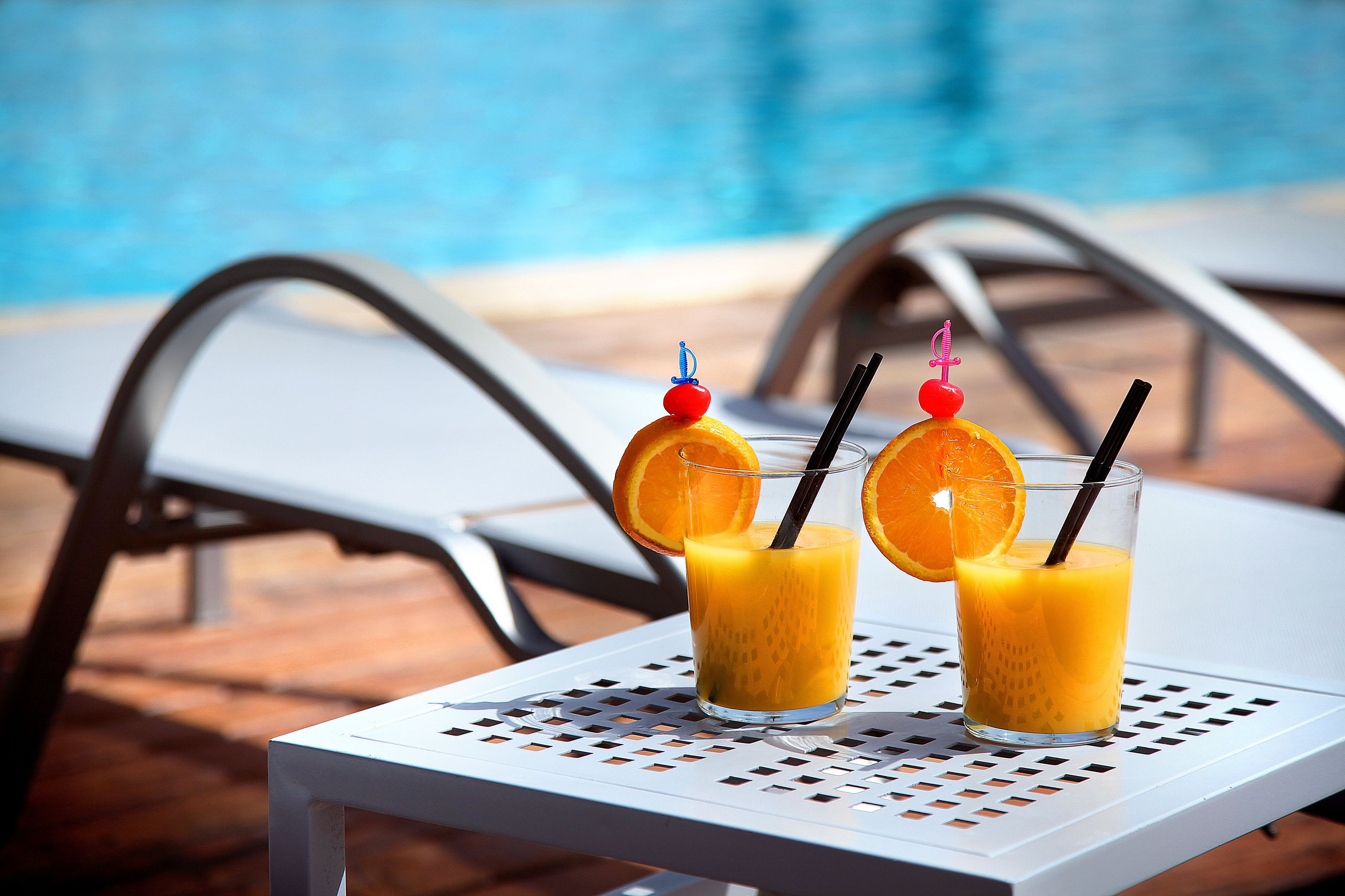 Dos vasos de jugo de naranja con pajitas y rodajas de naranja sobre una mesa blanca junto a una piscina.