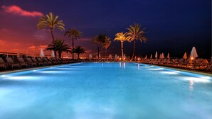 A swimming pool with lounge chairs and umbrellas, surrounded by palm trees, under a twilight sky.