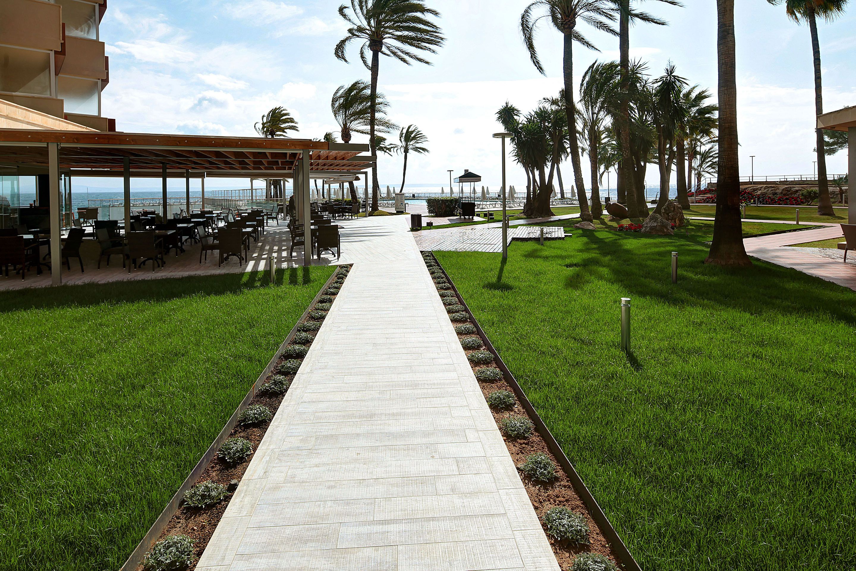 A pathway leading to outdoor seating with a view of the sea, surrounded by palm trees and a covered area with tables and chairs.