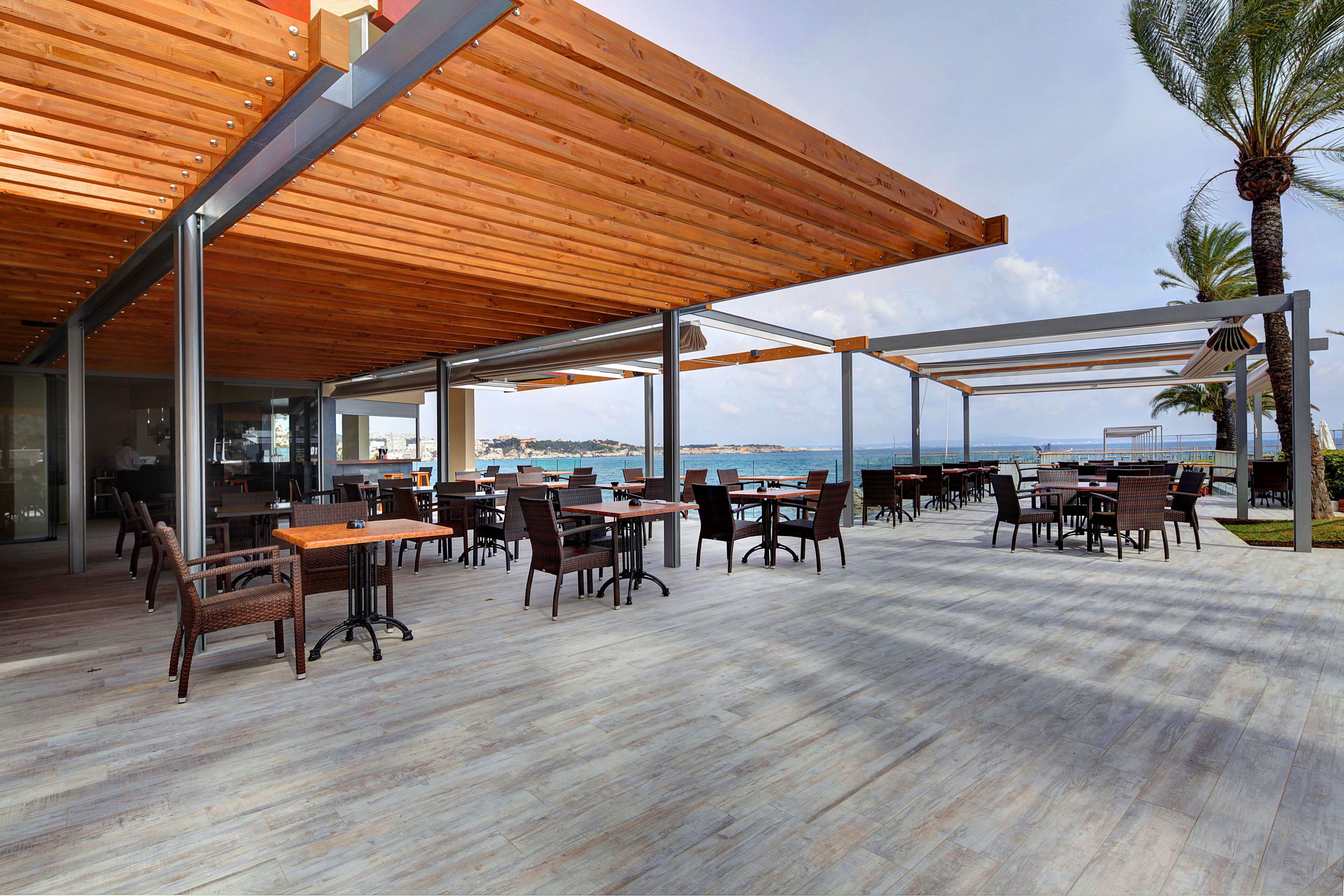 An outdoor dining area with wooden tables and chairs, a view of the sea, and a covered seating area.