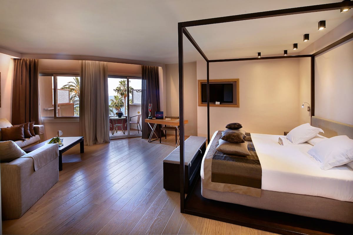 Une chambre d’hôtel avec un lit à baldaquin, un canapé, un bureau et un balcon offrant une vue sur les palmiers.