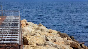 A rocky shoreline with metal sun loungers and a metal railing.
