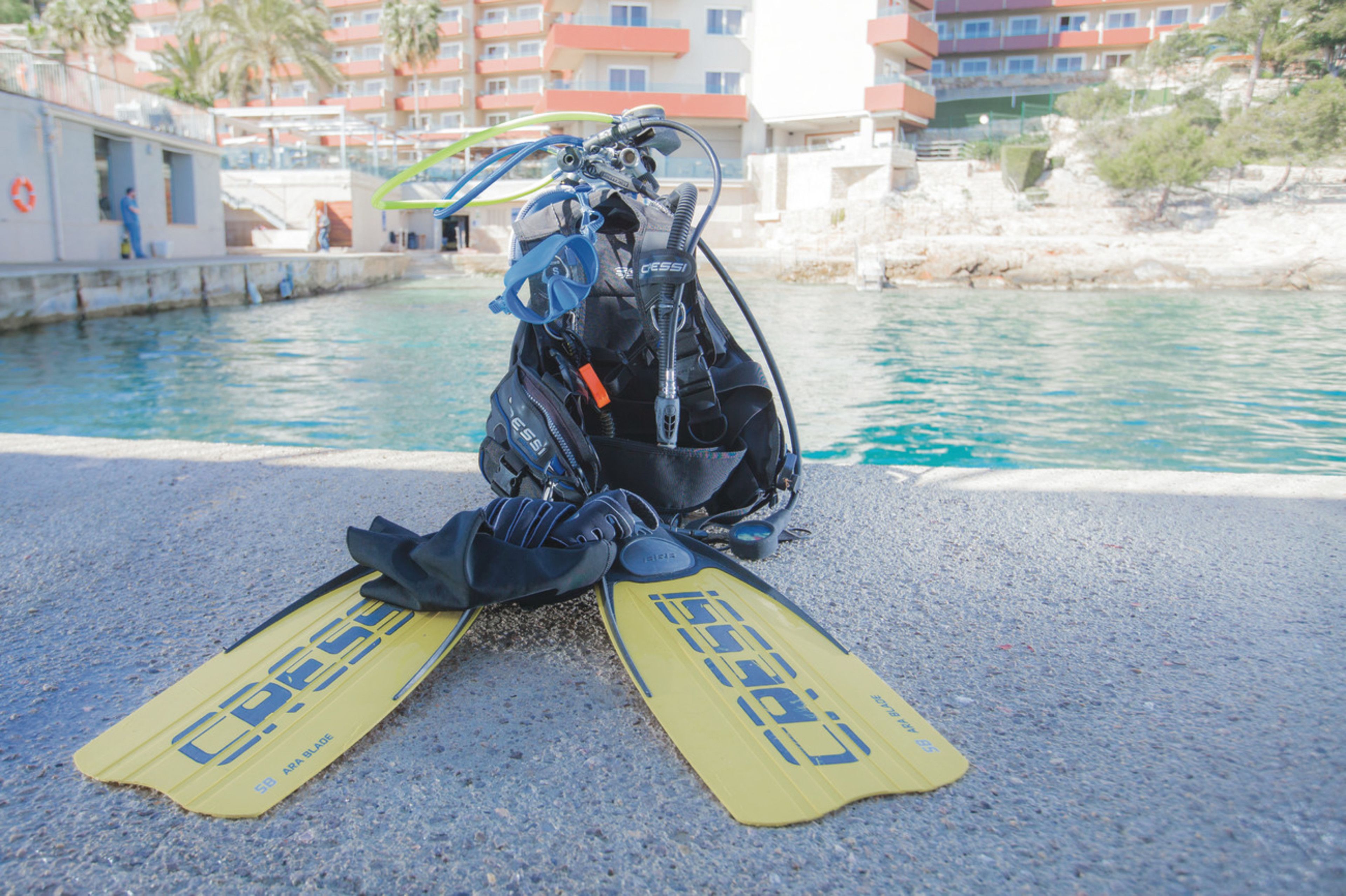 A scuba diving setup with fins and equipment on a concrete surface near a body of water, with buildings in the background.
