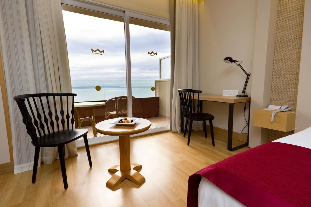 Et hotelværelse med balkon, et rundt træbord, to sorte stole, et skrivebord med lampe og en seng med rødt tæppe.
