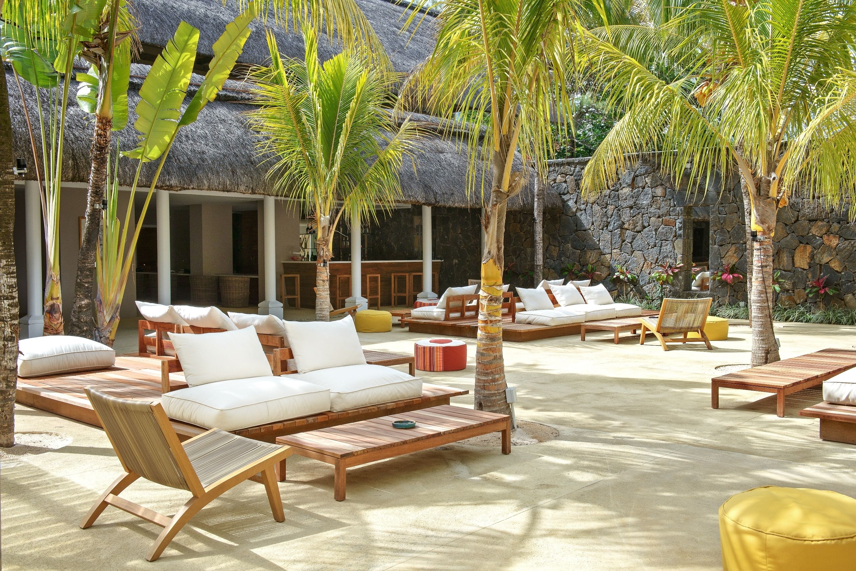 A tropical resort with thatched-roof buildings, wooden lounge areas, and palm trees.