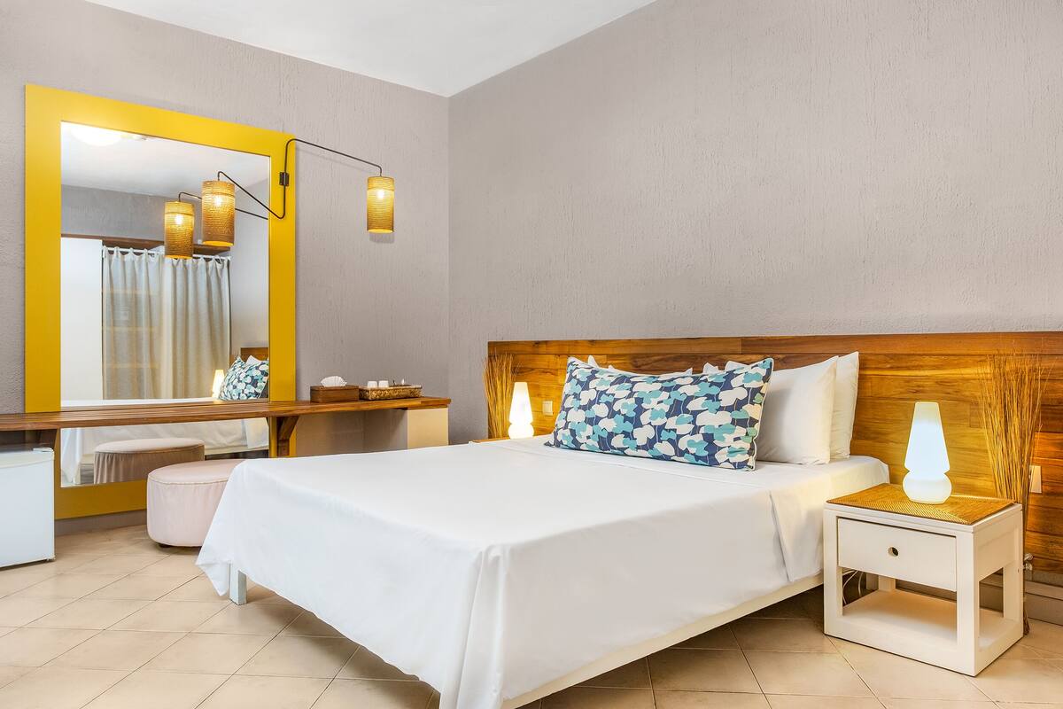 A neatly arranged hotel room with a large bed, a bedside table with a lamp, a nightstand, a mirror, and a wooden headboard.