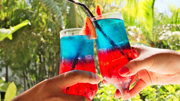 Two hands holding glasses of a blue and red drink with a straw and a slice of fruit, set against a tropical background with palm trees. - Friday Attitude (Trou d'Eau Douce)