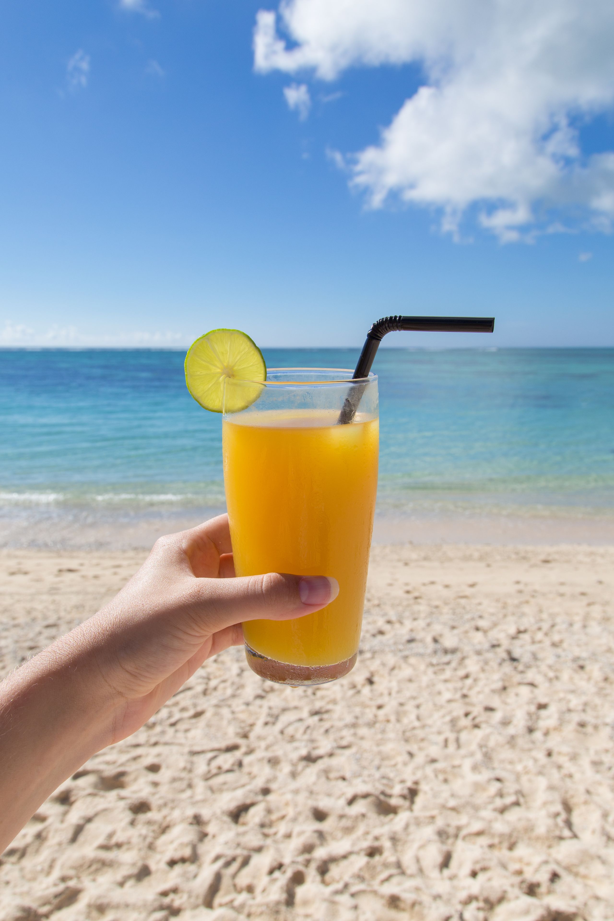 Une main tenant un verre de jus d’orange avec une paille et une tranche de citron vert, sur une plage où l’eau est d’un bleu limpide et le rivage est sablonneux.
