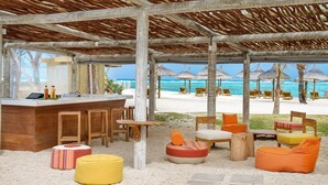 An outdoor seating area with wooden furniture and a bar, overlooking a beach with umbrellas.
