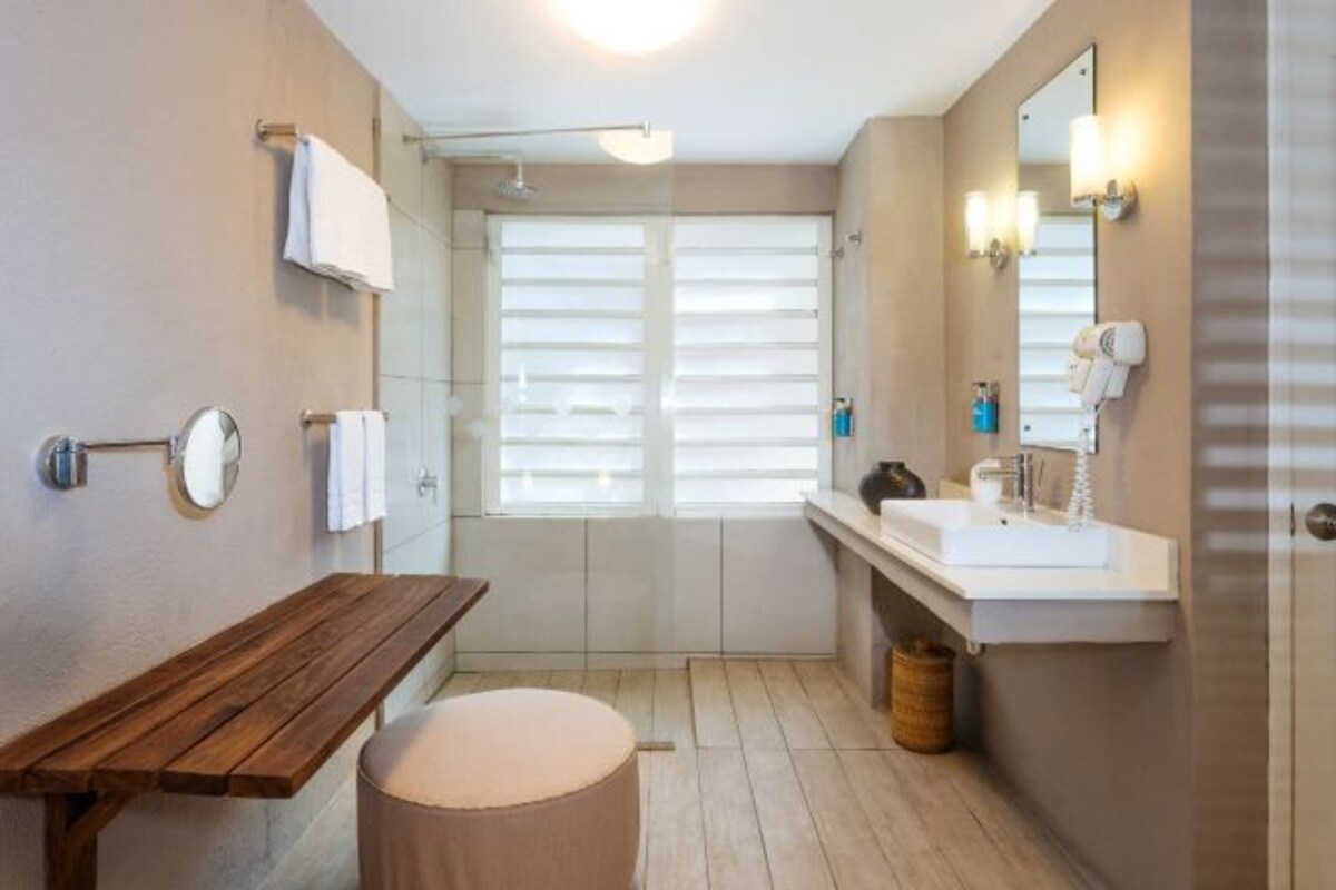 A modern bathroom with a wooden bench, a round stool, a sink with a mirror, and a shower area.