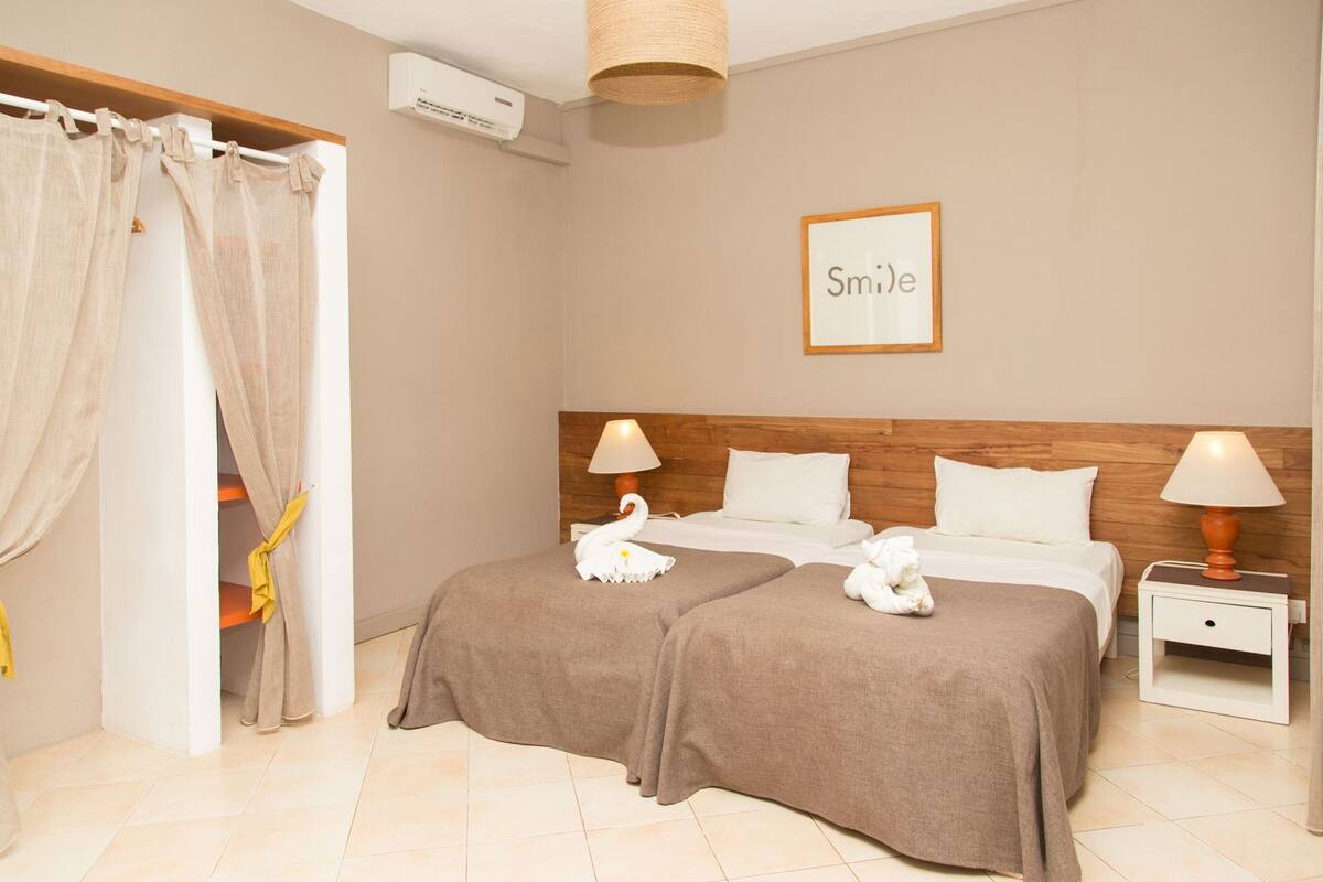 A hotel room with two beds, a wooden headboard, a wall-mounted air conditioner, a framed 'Smile' sign, and a small bedside table with a lamp.