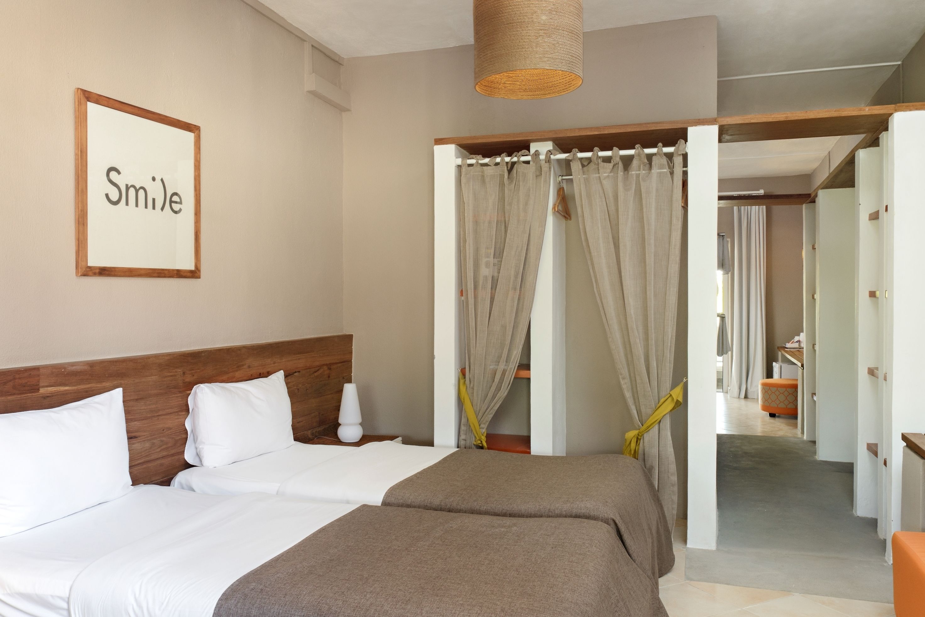 Een hotelkamer met een bed, een nachtkastje, een lamp, een bord met de tekst 'Smile' in een lijst aan de muur, en uitzicht op de gang.