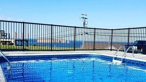A swimming pool with a metal fence, clear blue water, and a view of the ocean and hills in the background. - Hotel Motel Panorama de Perce (Perce)