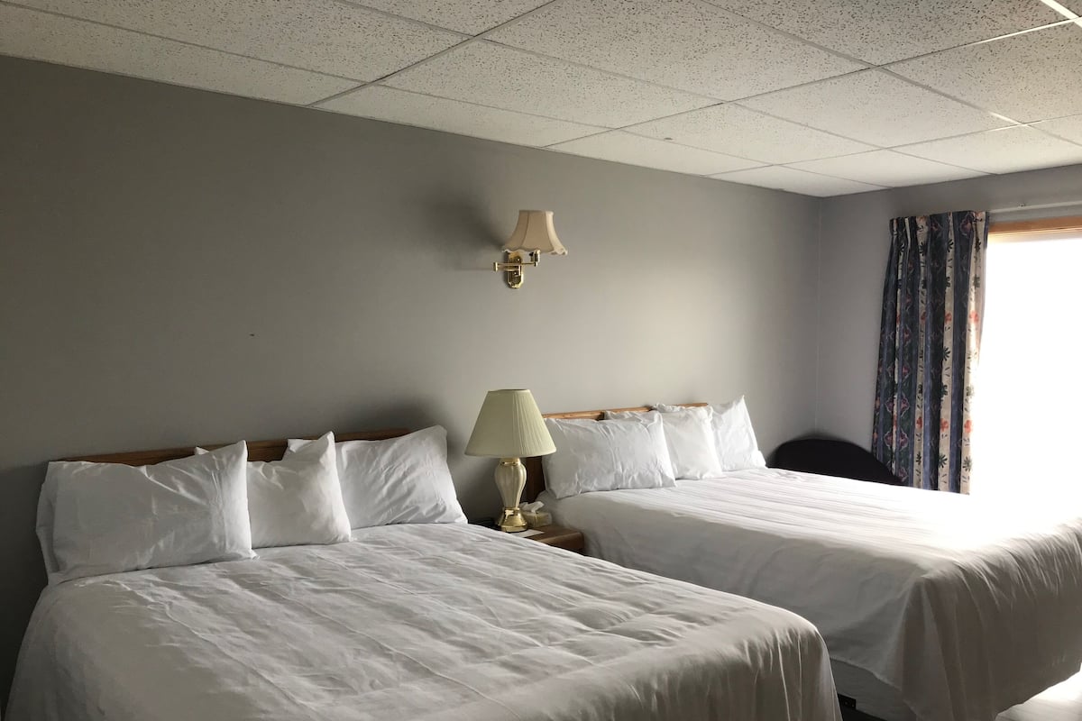 A hotel room with two single beds, a nightstand with a lamp, a wall-mounted light, and a window with curtains.