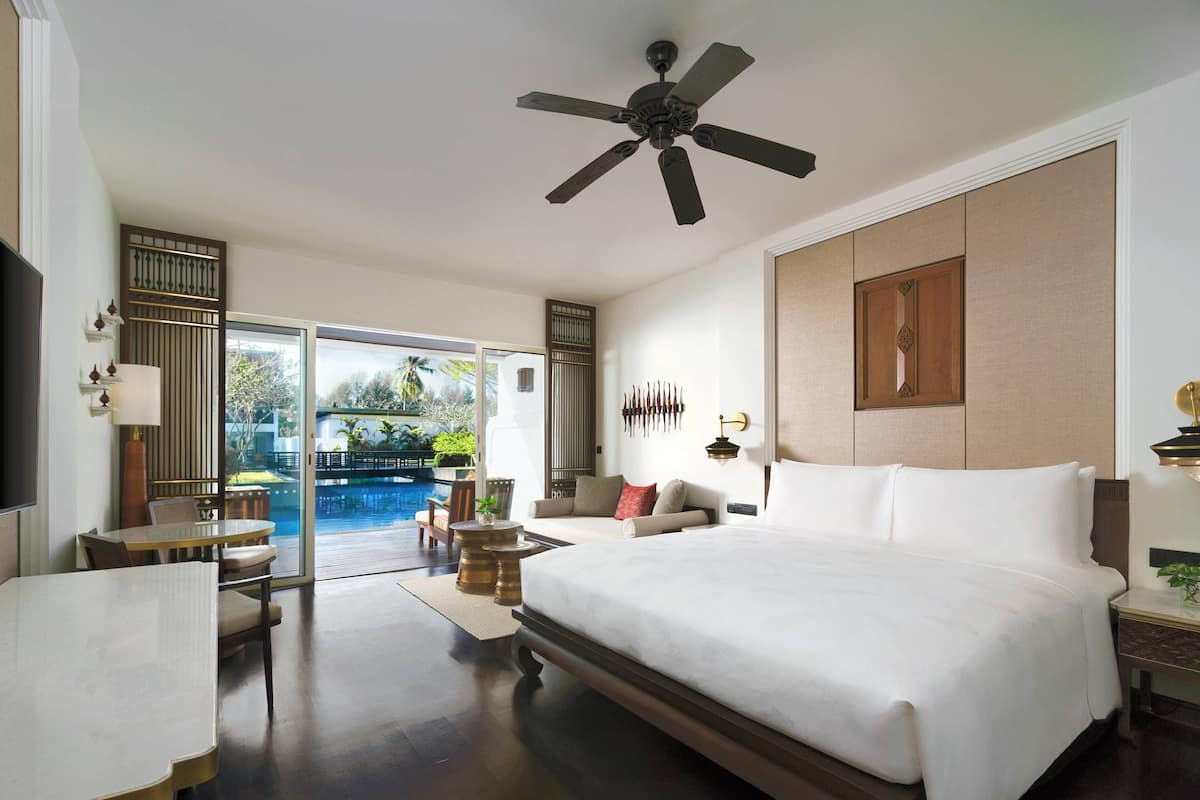 A spacious bedroom with a large bed, a ceiling fan, a dining area with a table and chairs, and a view of a swimming pool through glass doors.