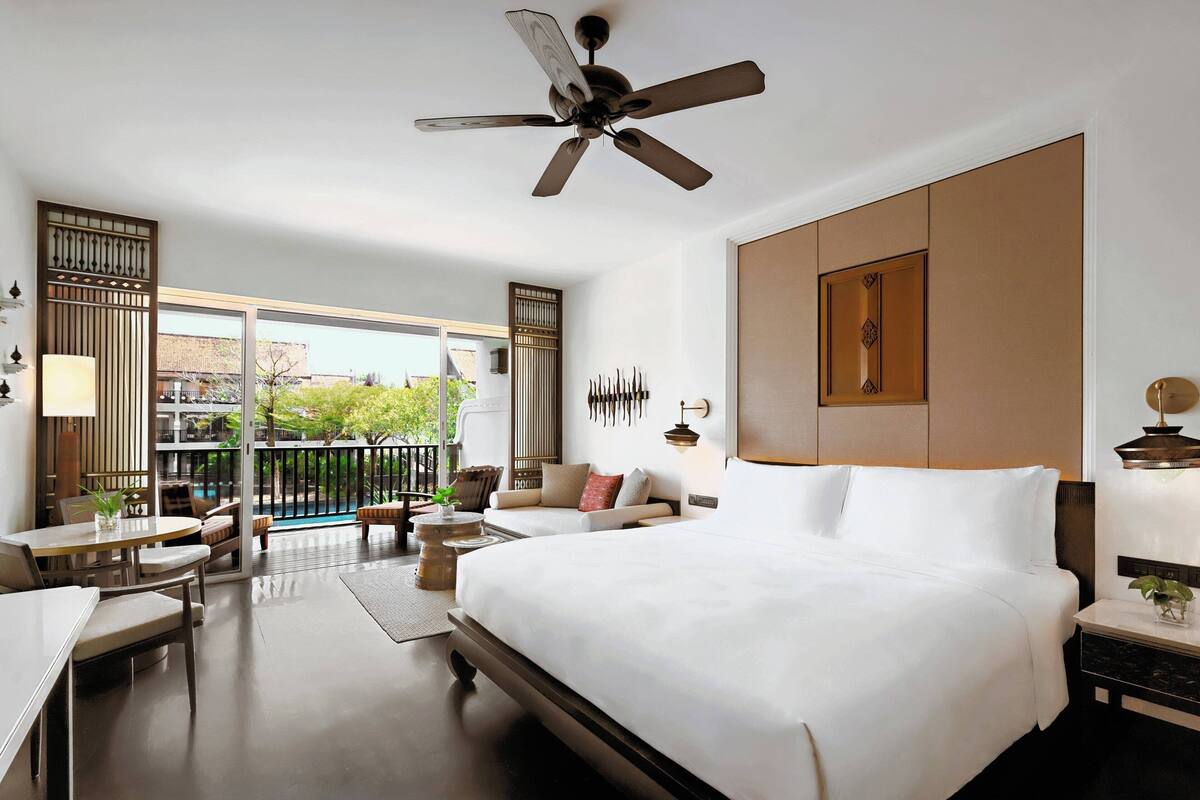 A hotel room with a large bed, a ceiling fan, a balcony with outdoor seating, and a view of the city.