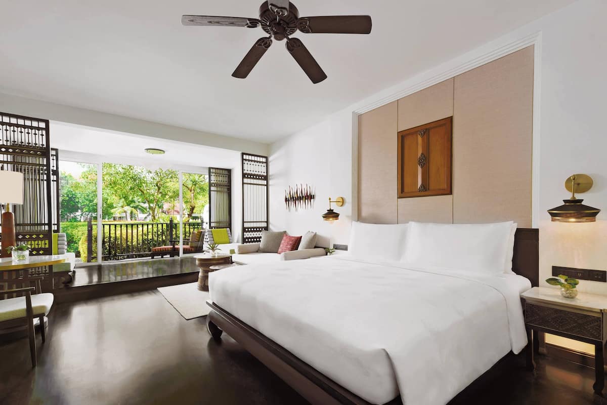 A spacious bedroom with a large bed, a ceiling fan, a nightstand, and a view of greenery outside.
