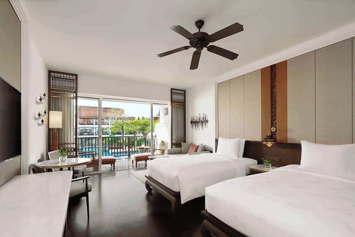 Ein Hotelzimmer mit zwei Betten, einem Deckenventilator, einem Schreibtisch mit Stuhl, einem kleinen Tisch und Blick auf einen Pool und Grünflächen.