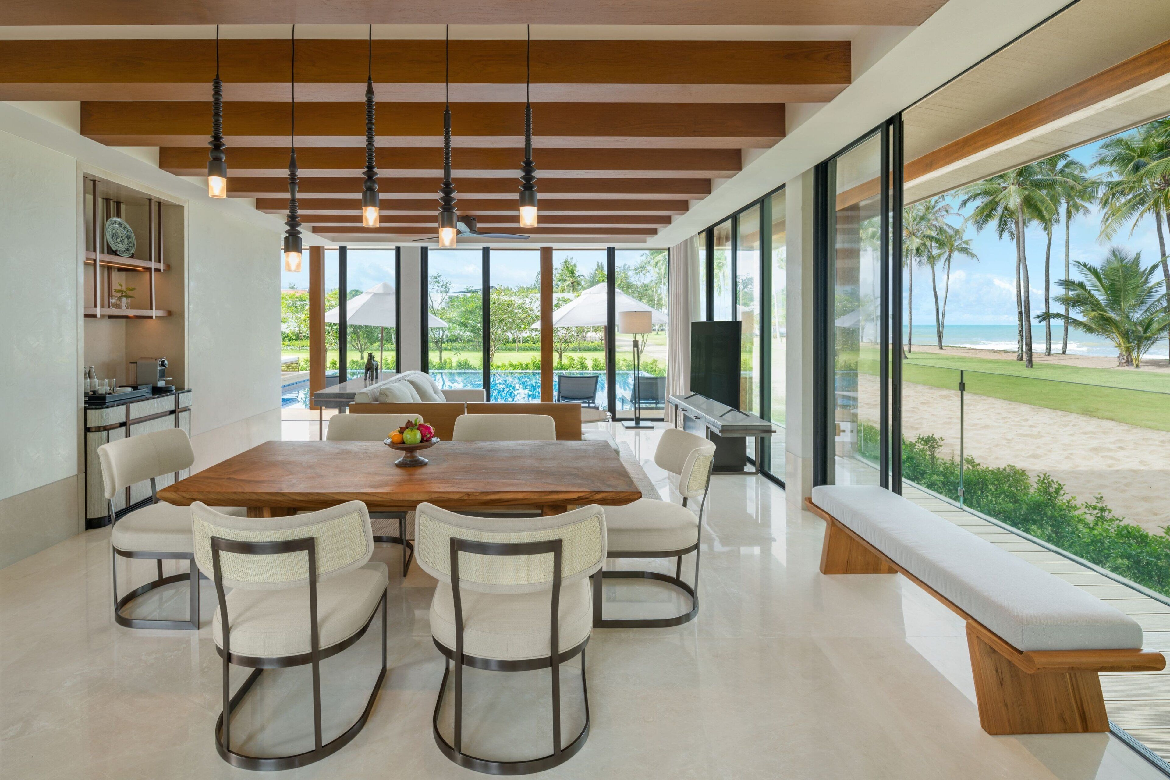 Un comedor moderno con una mesa y sillas de madera, ventanas amplias con vista a la playa y palmeras, y una zona de cocina con mostrador y armarios.