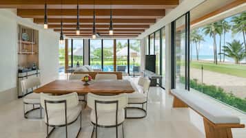 Un comedor moderno con una mesa y sillas de madera, ventanas amplias con vista a la playa y palmeras, y una zona de cocina con mostrador y armarios.