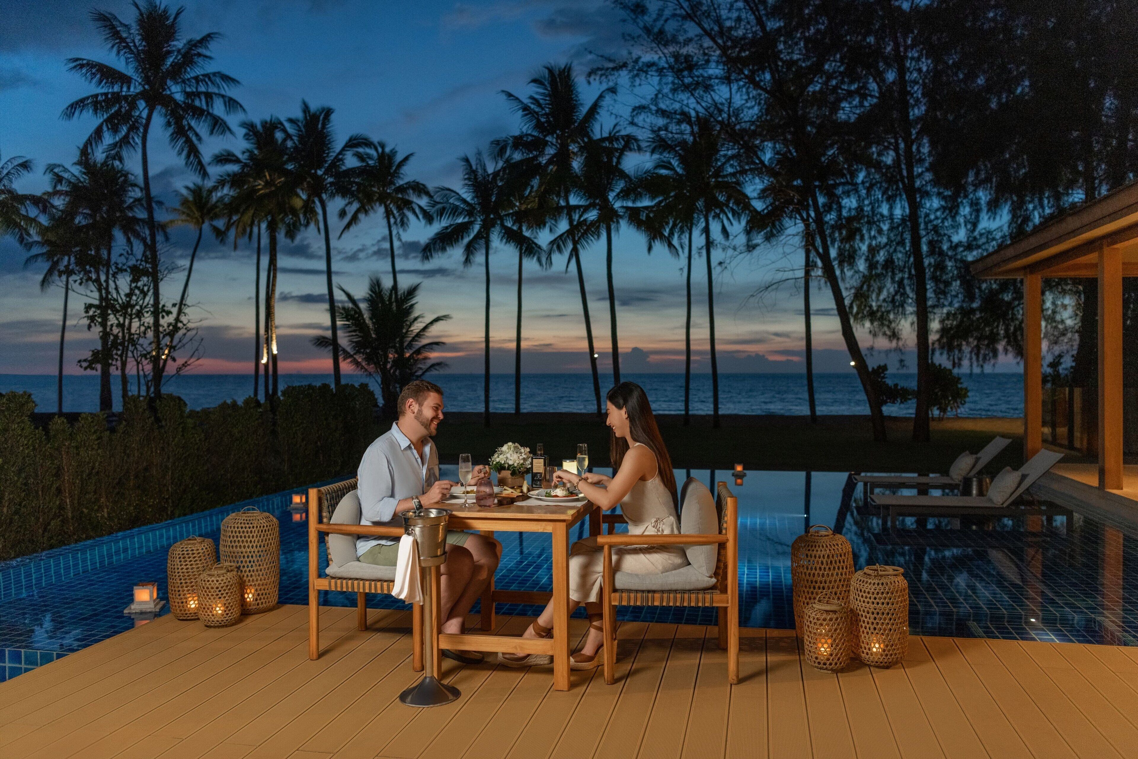 A couple dining at a wooden table by a pool with a view of palm trees and the ocean at dusk.