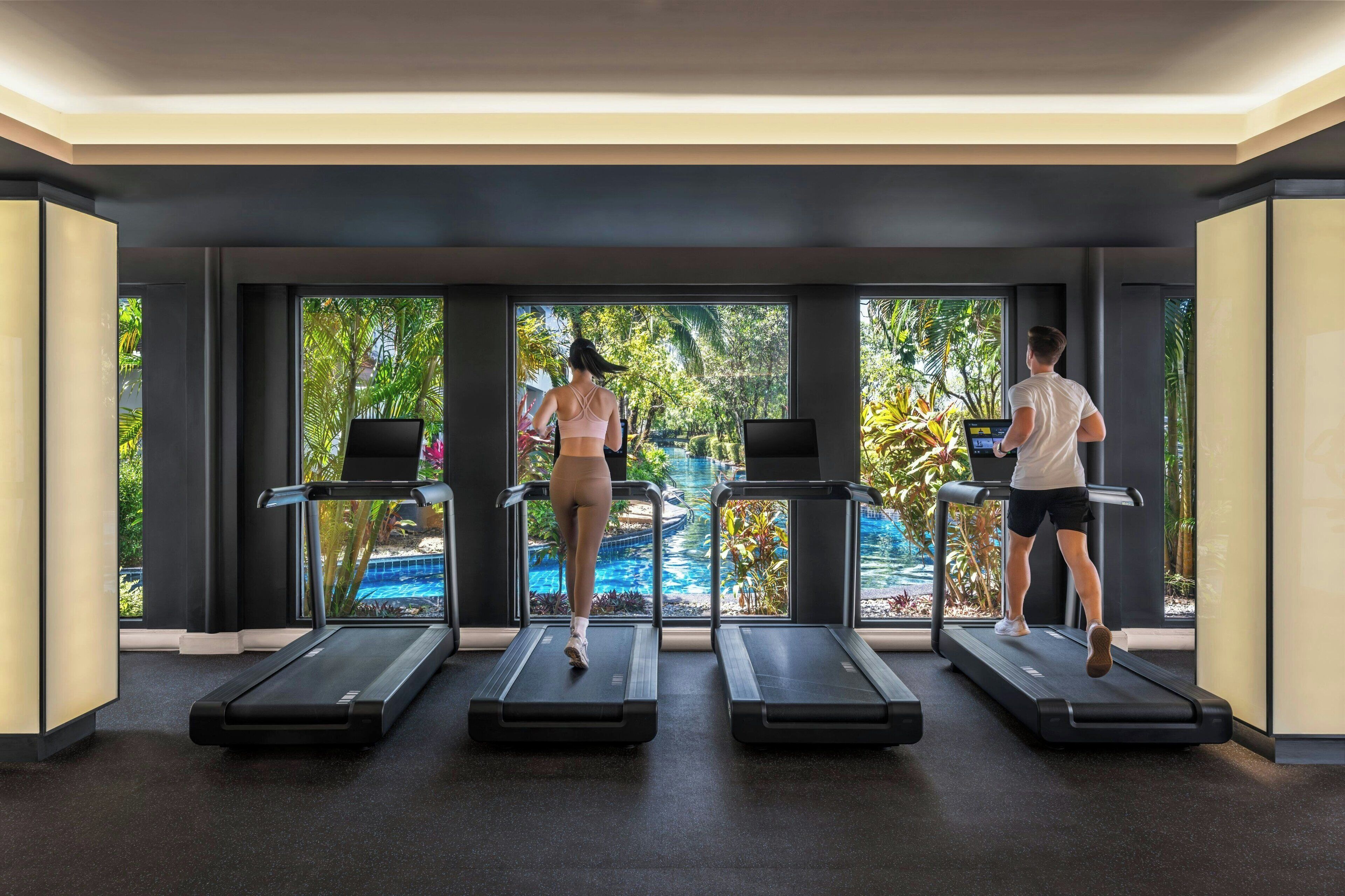 A gym with treadmills aligned in front of large windows with a view of a pool and greenery.