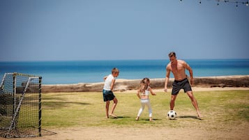 Partido de fútbol en la playa, con un arco cubierto por una red, un campo arenoso y un cielo azul despejado.