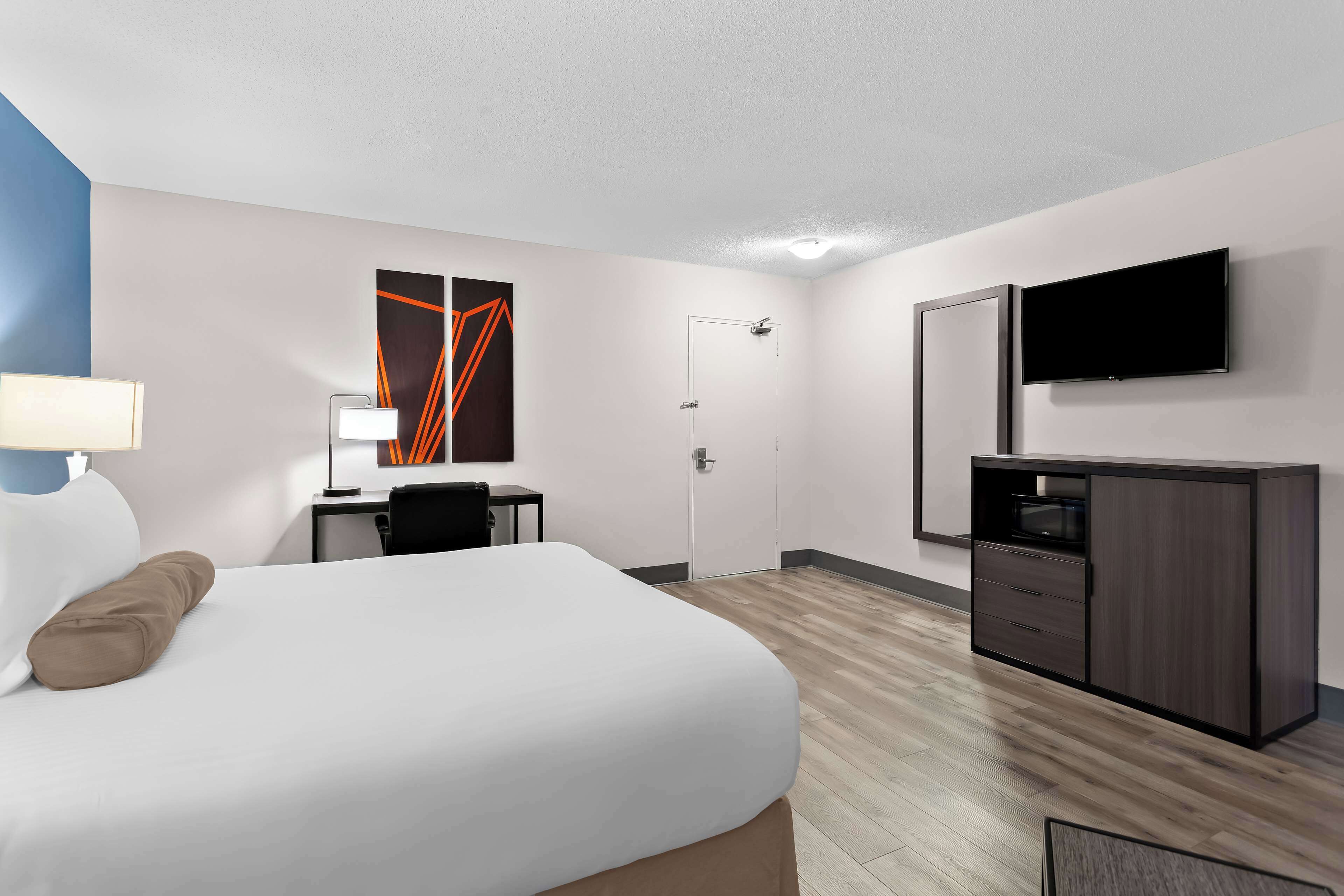 A hotel room with a bed, desk, chair, TV, wardrobe, and artwork on the wall.