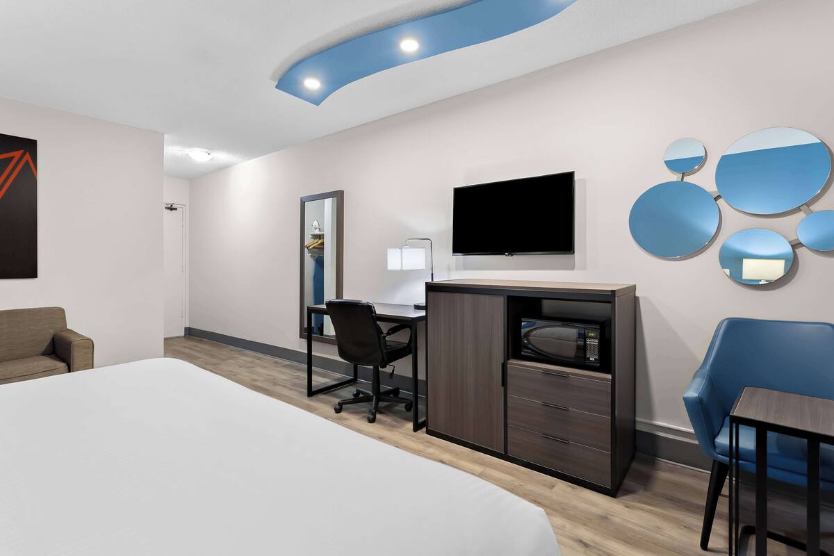 A modern hotel room with a large bed, a desk with a chair, a flat-screen TV, a mirror, and a blue armchair.