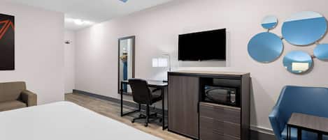 Une chambre d’hôtel moderne avec un grand lit, un bureau avec une chaise, un téléviseur à écran plat, un miroir et un fauteuil bleu.