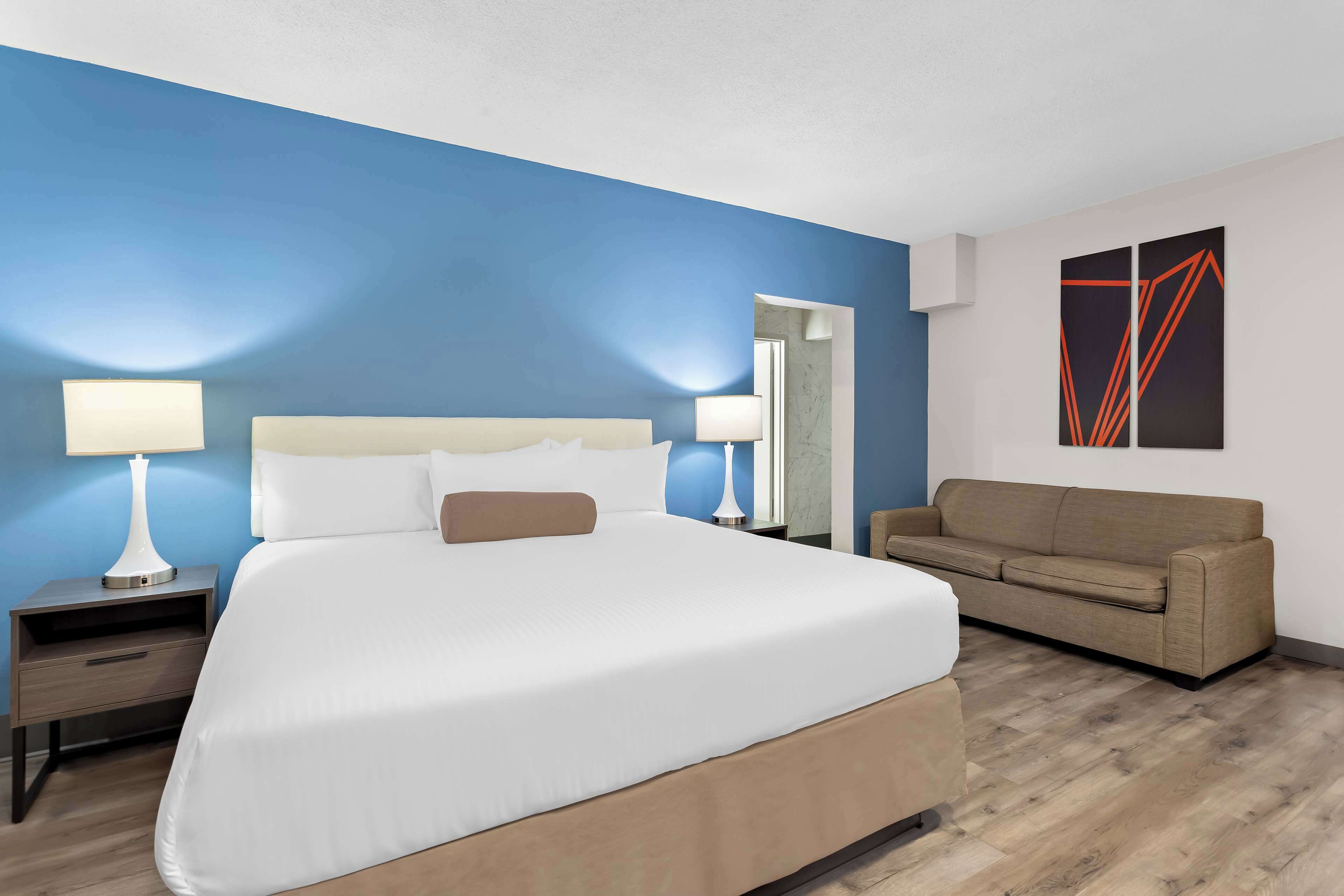 A hotel room with a large bed, two bedside tables with lamps, a beige sofa, and a blue accent wall.
